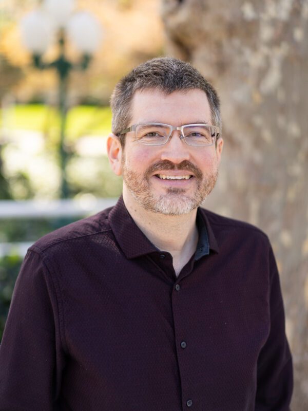 Outdoor photo of bearded man wearing glasses smiling in dress shirt with tree in background