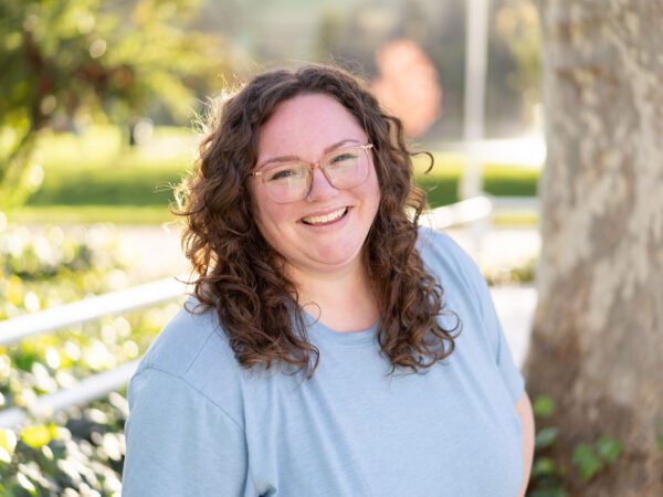 Outdoor photo of brunette woman wearing glasses smiling with blurry tree in background