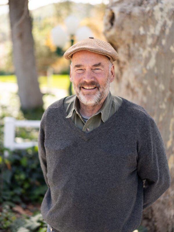Outdoor photo of bearded man smiling in sweater and hat with tree in background