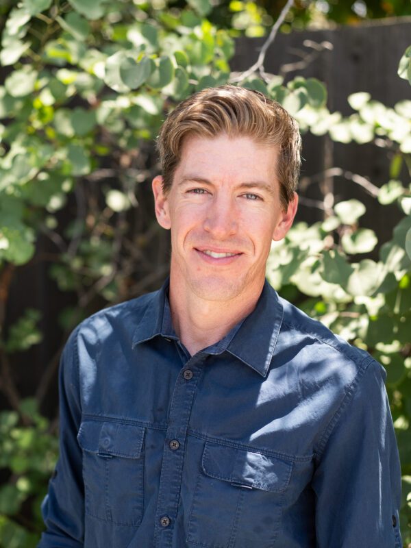 Outdoor photo of man smiling in dress shirt with sunlit leaves background