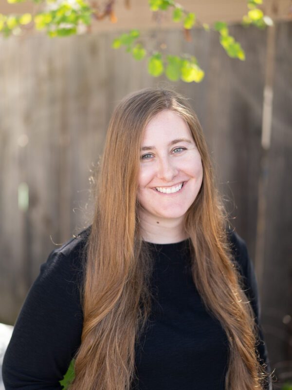 Outdoor photo of brown haired woman smiling with a black long sleeve and a blurry fence in background
