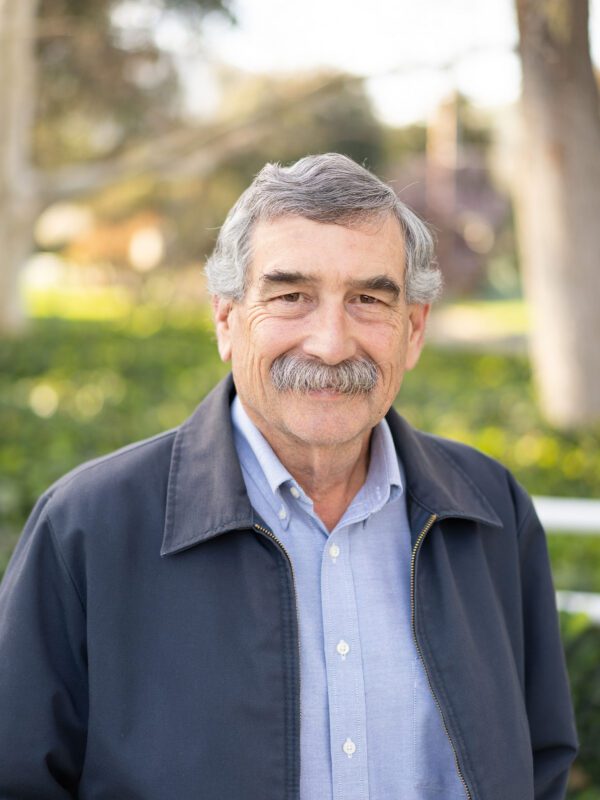 Outdoor photo of grey haired man with mustache smiling in coat with tree in background