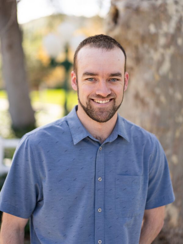 Outdoor photo of bearded man smiling in dress shirt with tree in background
