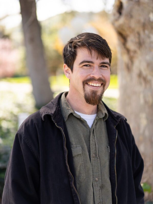 Outdoor photo of man with goatee smiling in coat with tree in background