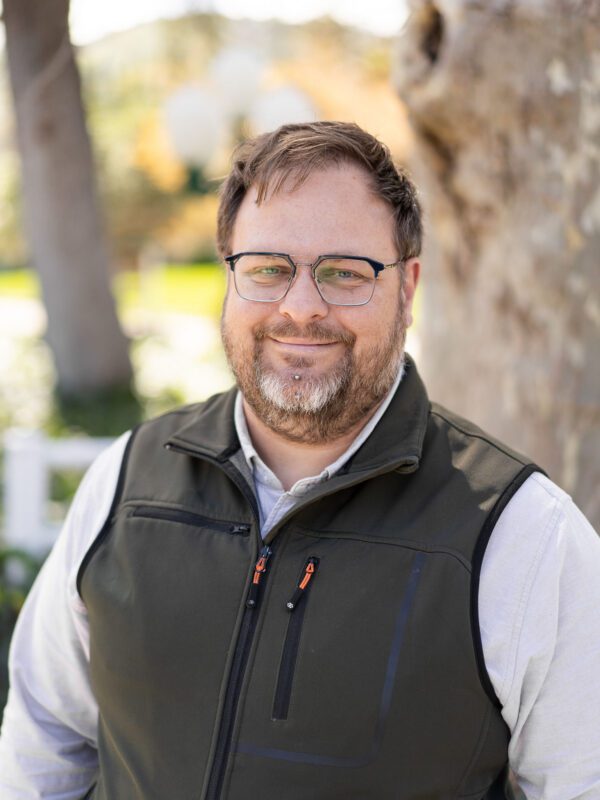 Outdoor photo of bearded man smiling in vest with tree in background