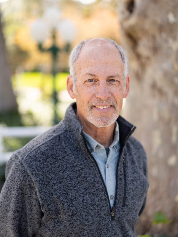 Outdoor photo of grey haired man with goatee smiling in coat and dress shirt with tree in background