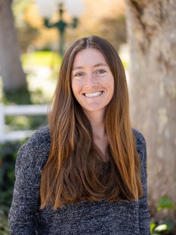 Outdoor photo of brown haired woman smiling with a grey sweater and a blurry tree in background