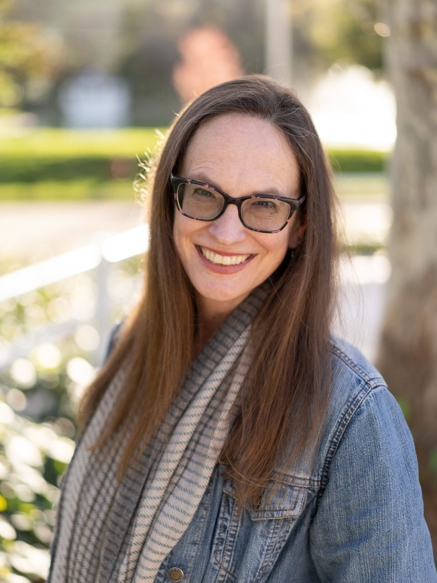 Outdoor photo of brunette woman wearing glasses, scarf and denim jacket smiling with blurry tree in background