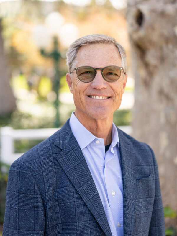 Outdoor photo of grey haired man with glasses smiling in coat and dress shirt with tree in background