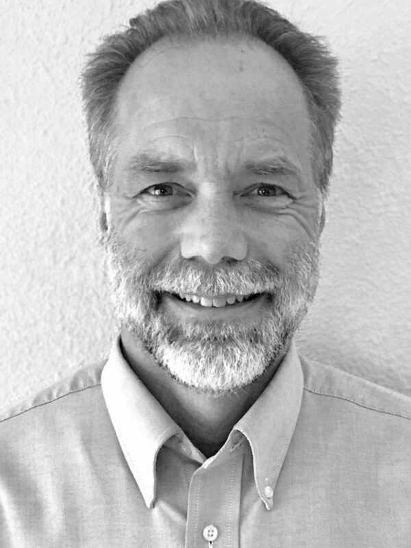 Close up black and white photo of grey haired bearded man smiling wearing a dress shirt.