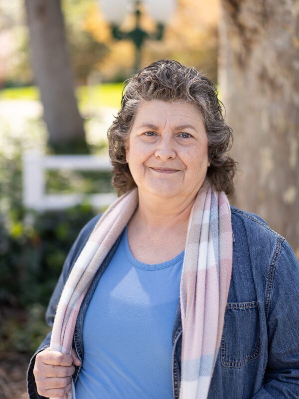 Outdoor photo of grey haired woman smiling wearing scarf and denim jacket with blurry tree in background