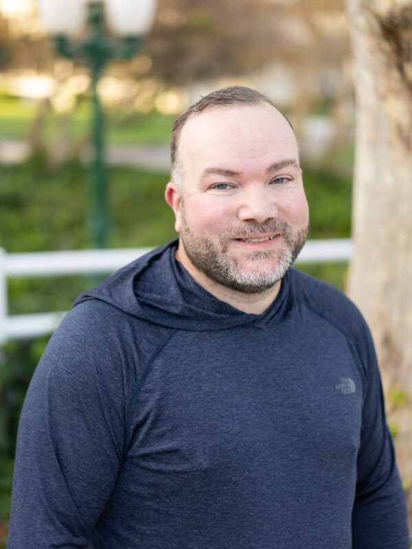 Outdoor photo of man smiling in grey long sleeve shirt with tree in background
