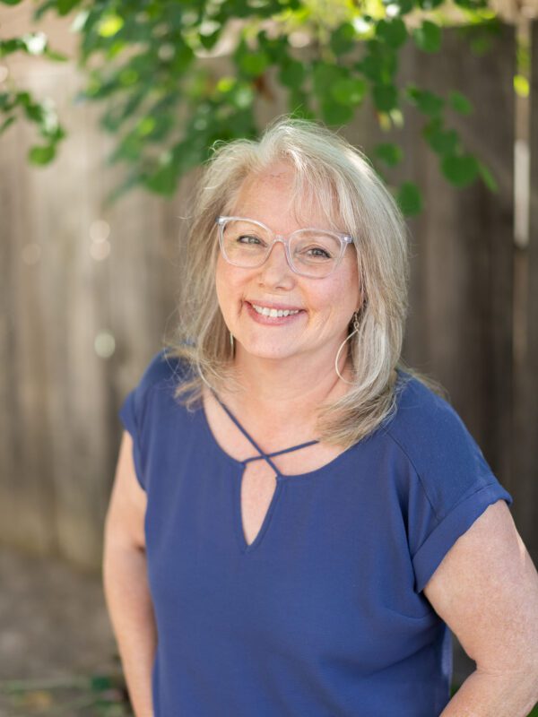 Outdoor photo of grey haired woman smiling wearing a blue top with blurry fence and leaves in background