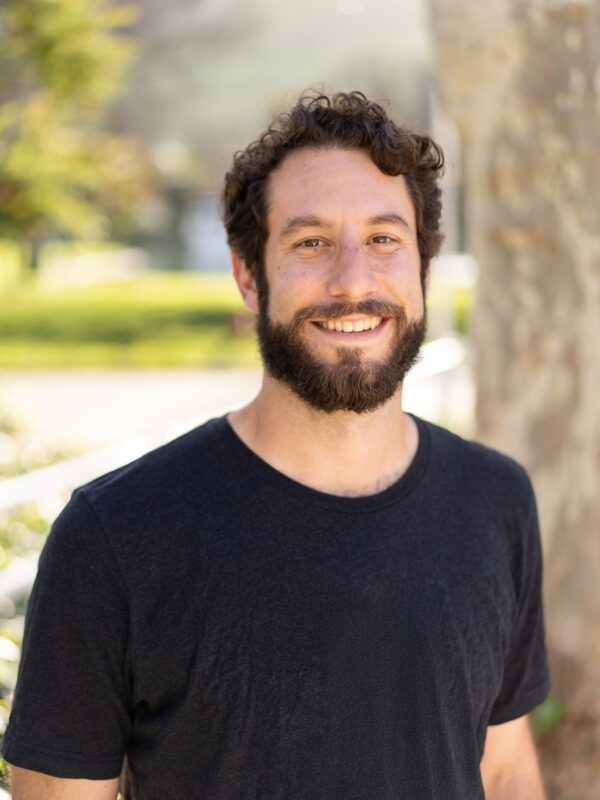Outdoor photo of bearded man smiling in black t shirt with tree in background
