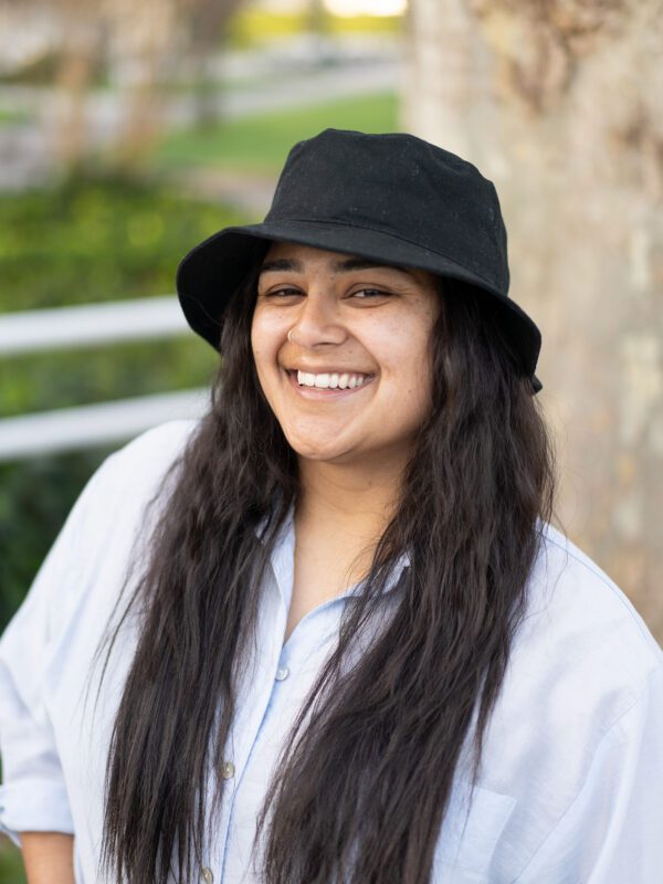 Outdoor photo of brunette woman wearing a hat and dress shirt smiling with blurry tree in background