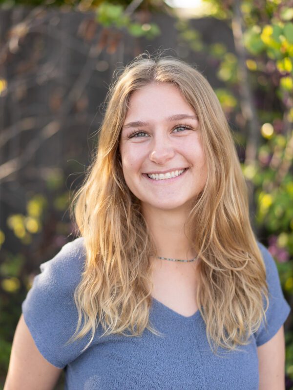 Outdoor photo of blonde woman smiling with blurry leaves in background