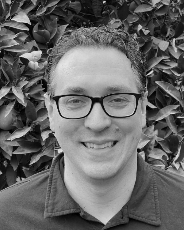 Close up black and white photo of dark haired man smiling wearing a dress shirt in front of leaves.
