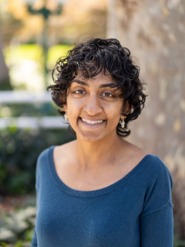 Outdoor photo of curlly haired woman smiling with a blue top and a blurry tree in background