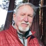 man with grey hair and white goatee smiling wearing red jacket with redwood trees in background