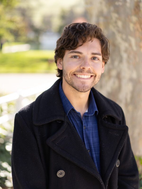 Outdoor photo of man smiling in coat with tree in background